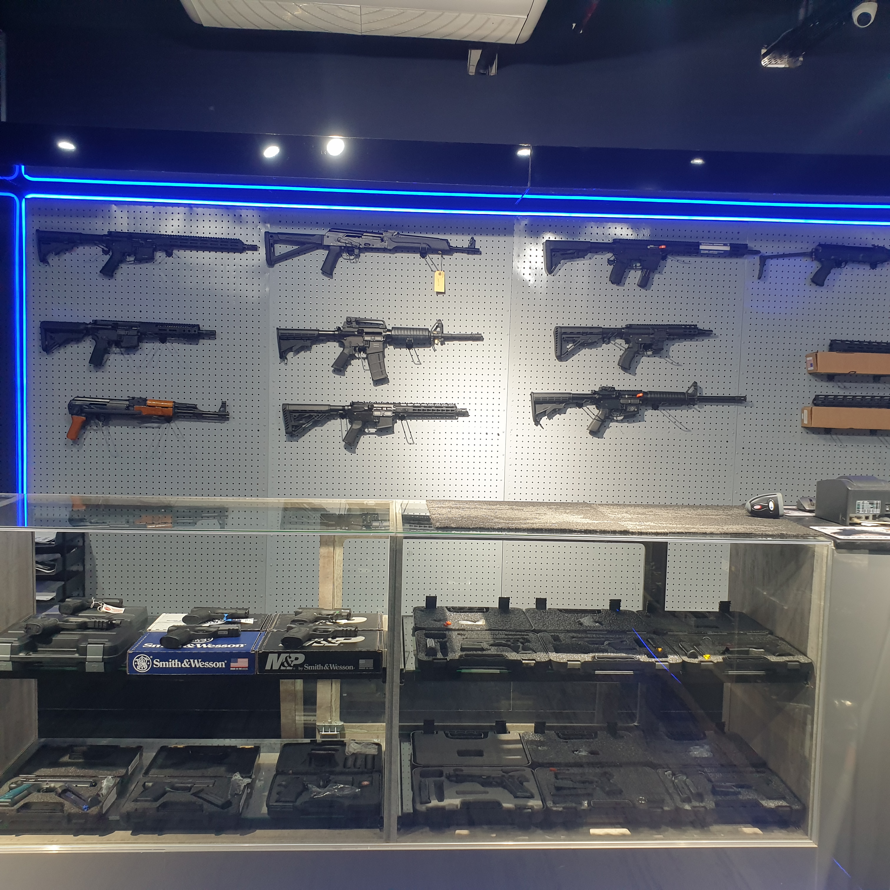 Pegboard rifle wall in blue LED light above a glass counter of Smith & Wesson boxes and pistol cases