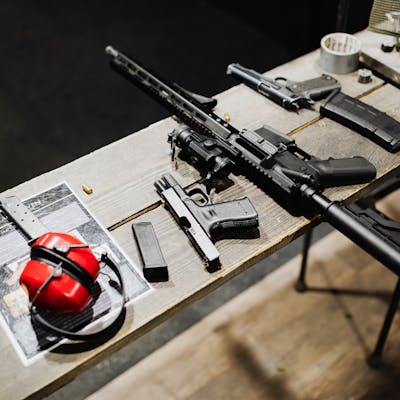 A variety of guns and accessories displayed on a wooden table indoors, featuring ear protection.